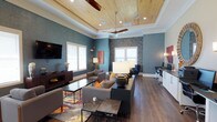 This single-floor space presents a thoughtfully designed lounge or common area that blends contemporary style with warm, inviting elements. The standout architectural feature is the coffered tray ceiling, finished with natural wood tongue-and-groove paneling and complemented by recessed lighting and dual ceiling fans, adding both visual interest and comfort to the room.

The walls are dressed in a muted teal-blue textured finish, punctuated by a bold, mosaic-style accent wall that serves as a focal point and adds depth to the space. Rich hardwood flooring runs throughout, grounding the room with warmth and continuity.

The lounge is furnished with generous, modern seating arrangements, including plush gray sofas accented with mustard throw pillows, paired with sleek coffee tables and a vibrant area rug that introduces color and pattern. A wall-mounted flat-screen television is positioned near a dark wood console, enhancing the room's entertainment functionality.

Toward the perimeter, a workstation area with white cabinetry provides a practical counterpoint to the relaxed seating zones. Multiple windows dressed with white blinds allow natural light to fill the space. Overall, the room exudes a polished, resort-inspired ambiance ideal for gathering and relaxation.