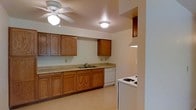 This single-floor space includes a kitchen designed for everyday functionality. The kitchen features warm-toned, raised-panel wood cabinetry that lines the walls, providing ample upper and lower storage. A continuous countertop runs the length of the cabinetry, offering generous prep space. The double-basin sink is centrally positioned beneath the upper cabinets, accompanied by a white dishwasher. A coil-burner electric cooktop and a white refrigerator are positioned along an adjacent wall, completing the suite of appliances. The light-toned vinyl flooring reflects the overhead lighting, contributing to a bright atmosphere. Illumination is provided by a central ceiling fan with an integrated light fixture, as well as a flush-mount ceiling light, ensuring the space is well-lit throughout. The neutral wall tones complement the wood cabinetry and create a clean, understated backdrop. The layout is straightforward and efficient, with clear sightlines throughout the kitchen, making it a practical and accessible space for daily use.