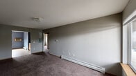 Located in Marion, Iowa, this single-floor unit offers a practical and straightforward layout across several connected rooms. The living area is finished with neutral gray walls and wall-to-wall carpet, creating a simple, understated atmosphere. A baseboard heating system runs along the exterior walls, providing efficient warmth throughout. Windows in the living and bedroom areas allow natural light to filter in and offer views of the surrounding neighborhood and greenery.

The kitchen is a functional space featuring warm wood cabinetry both above and below the countertops, a double-basin sink, a white electric range, and a refrigerator. The kitchen is finished with tile flooring and accented by deep blue walls, giving it a distinctive look compared to the rest of the unit. Ample cabinet storage is available for everyday needs.

The bedrooms are carpeted and neutral in tone, each offering adequate space and window access. One bedroom includes a wall-mounted air conditioning unit alongside baseboard heating, ensuring year-round climate control. A closet area with a clothes rod is also present within the unit.

Overall, this Marion residence presents a clean, no-frills living environment with all essential amenities in place, suited for comfortable everyday living.