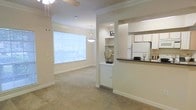 This single-floor residential space offers a warm and inviting open-concept layout that connects the living, dining, and kitchen areas into one cohesive flow. The interior is finished in soft, neutral tones throughout, with light beige walls and plush wall-to-wall carpeting that contribute to a comfortable and welcoming atmosphere. A large window in the dining area floods the space with abundant natural light, creating an airy and bright environment. A stylish pendant light hangs above, adding a refined decorative touch to the dining zone. Crown molding along the ceilings lends an element of classic architectural elegance, while a ceiling fan in the living area adds both function and subtle design interest. The kitchen is accessible via a pass-through opening framed by a dark countertop bar, providing a practical and social connection between the cooking and living spaces. White cabinetry and a full-size refrigerator are visible within the kitchen, offering a clean and functional layout. A built-in shelving niche near the kitchen pass-through provides a charming display area for décor. Overall, the space balances understated design with practical features, making it a comfortable and well-appointed single-level home.