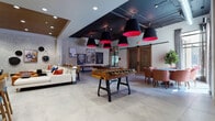 Located at 100 Union St in Elizabeth, NJ, this dynamic single-floor community lounge is designed to impress with its eclectic mix of industrial and contemporary aesthetics. The space features exposed whitewashed brick walls adorned with curated artwork, warm wood-slat ceiling accents, and dramatic black pendant lights with bold red interiors that draw the eye upward. Large-format light-toned tile flooring runs throughout, lending a clean, cohesive look to the expansive open layout. A central foosball table anchors the main gathering area, while a dedicated seating zone offers generous sectional sofas with vibrant accent pillows, a tufted leather ottoman, and eclectic wall art that adds personality and depth. A separate lounge nook features a reclaimed wood accent wall, wall-mounted televisions, and stylish dining seating—ideal for casual gatherings or screen-side socializing. Rustic sliding barn doors provide a striking architectural contrast against the more industrial exposed ceiling elements and concrete structural columns. Additional seating vignettes with accent chairs are thoughtfully positioned throughout, creating intimate conversation areas within the larger open plan. A custom artwork panel celebrating the city of Elizabeth greets visitors near the entrance, reinforcing a strong sense of local identity. Natural light filters in through windows, complementing the layered ambient and accent lighting to create a welcoming, vibrant atmosphere throughout the day.