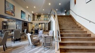 The club room is a thoughtfully designed communal space that combines modern sophistication with warm, inviting elements. The double-height ceiling creates an impressive sense of openness, drawing the eye upward and amplifying the overall sense of scale throughout the room. A prominent wood-tread staircase with sleek glass and metal railings serves as a focal architectural element, adding both visual interest and a refined, contemporary character.

The main lounge area is furnished with plush, neutral-toned seating arrangements, including upholstered armchairs and accent chairs positioned around low tables — ideal for conversation and relaxation. Large-format tile flooring with a wood-look finish runs throughout, contributing to the modern aesthetic while remaining practical. Recessed lighting throughout the ceiling provides well-distributed illumination, complementing the ambient glow of accent lamps.

A stone-clad feature wall with an integrated fireplace anchors one side of the space, adding warmth and a luxurious touch. Colorful abstract artwork is displayed on the walls, adding personality and visual vibrancy. An upper mezzanine level, visible behind glass railings, extends the usable area and adds architectural depth to this well-appointed gathering space.