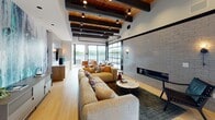 The Vestry Club Room in Montclair, NJ is a thoughtfully designed single-floor communal space that blends industrial aesthetics with modern comfort. The architecture is defined by exposed dark wood ceiling beams set against warm wood planking, complemented by black steel accents and track lighting that add a contemporary edge throughout.

The space is anchored by a generous lounge area featuring a large sectional sofa and accent chairs arranged around a linear fireplace set into a stacked stone wall. Adjacent to the lounge, a bold abstract mural in teal and charcoal tones creates a dramatic focal point, paired with a mounted flat-screen television and a sleek built-in media console with subway tile surround.

A dedicated game area includes a full-size billiard table positioned beneath the soaring beamed ceiling, with striking floor-to-ceiling glass garage-style doors that open to a terrace overlooking the surrounding landscape. These oversized doors flood the interior with natural light and provide seamless indoor-outdoor connectivity.

Light hardwood flooring runs throughout, tying together the various zones of the space. The overall ambiance is sophisticated yet relaxed, making The Vestry Club Room a versatile venue for both casual gatherings and social events.