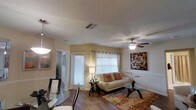 This single-floor residence offers a thoughtfully designed open-concept layout that seamlessly connects the living and dining areas, creating a cohesive and functional living environment. The living room is a comfortable and inviting space, anchored by rich hardwood-style flooring that extends throughout the main areas and complemented by warm yellow-toned walls that lend a welcoming ambiance. A ceiling fan with a light fixture serves as a practical centerpiece overhead, while multiple windows dressed with white horizontal blinds and sheer curtains allow generous natural light to permeate the room. The living area is well-suited for relaxation and entertaining, with ample space for a sofa, accent chairs, and a glass-top coffee table. The adjacent dining area features a glass-top dining table with dark-framed chairs beneath a hanging light fixture, providing an elegant and intimate setting for meals. Classic wainscoting detail along the lower walls adds a touch of architectural character to the space. Wall art and decorative accents throughout contribute to the polished, move-in-ready aesthetic. A hallway visible from the main living area suggests additional rooms beyond, completing this well-appointed single-level home.