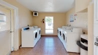 This single-floor communal laundry facility is designed to serve a residential community with efficiency and practicality in mind. The main laundry room features a generous arrangement of front-loading and top-loading washing machines lining both sides of the space, complemented by a bank of stacked commercial dryers along one wall, providing ample capacity for multiple users simultaneously. The room is finished with durable tile flooring in a warm earth tone, and the walls are painted in a neutral beige that lends the space a clean, welcoming feel. A large window centered on one wall floods the room with natural light and offers a view of the surrounding outdoor area. An HVAC unit mounted near the ceiling ensures comfortable temperatures year-round, while a mounted fire extinguisher box underscores the facility's attention to safety. An adjacent room serves as a utility or folding area, featuring the same tile flooring and warm wall tones, with direct exterior door access for added convenience. A simple table with hairpin legs provides a practical surface for sorting and folding laundry. Windows in this secondary space allow additional natural light to filter through, making the overall facility bright and functional throughout.