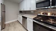 This single-floor, 2-bedroom, 2-bathroom residence offers a clean, modern aesthetic defined by neutral gray walls, white trim, and a cohesive design language throughout. The open-concept living and dining area is anchored by wood-look luxury vinyl plank flooring, a contemporary track lighting fixture, and a small chandelier that adds a refined touch near the entry. A pass-through peninsula connects the living space to the kitchen, maintaining an open, airy feel. Natural light enters through windows overlooking what appears to be a balcony or outdoor area.

The kitchen is well-equipped with white shaker-style cabinetry, granite-look countertops, stainless steel appliances including a dishwasher, range, and microwave, and a striking herringbone tile backsplash that adds visual interest. Wood-look flooring continues through this space seamlessly.

Both bedrooms are carpeted and finished in soft gray tones, offering a comfortable, quiet atmosphere. One bedroom features a sliding closet with ample storage, while windows with horizontal blinds bring in natural light. Each room provides a functional, versatile layout.

The bathrooms are updated with dark wood-tone vanities, integrated white sinks, backlit mirrors, brushed nickel hardware, and full tub-shower combinations with clean white surrounds and wood-look flooring.