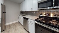 This single-floor, 2-bedroom, 2-bathroom residence offers a clean, modern aesthetic defined by neutral gray walls, white trim, and a cohesive design language throughout. The open-concept living and dining area is anchored by wood-look luxury vinyl plank flooring, a contemporary track lighting fixture, and a small chandelier that adds a refined touch near the entry. A pass-through peninsula connects the living space to the kitchen, maintaining an open, airy feel. Natural light enters through windows overlooking what appears to be a balcony or outdoor area.

The kitchen is well-equipped with white shaker-style cabinetry, granite-look countertops, stainless steel appliances including a dishwasher, range, and microwave, and a striking herringbone tile backsplash that adds visual interest. Wood-look flooring continues through this space seamlessly.

Both bedrooms are carpeted and finished in soft gray tones, offering a comfortable, quiet atmosphere. One bedroom features a sliding closet with ample storage, while windows with horizontal blinds bring in natural light. Each room provides a functional, versatile layout.

The bathrooms are updated with dark wood-tone vanities, integrated white sinks, backlit mirrors, brushed nickel hardware, and full tub-shower combinations with clean white surrounds and wood-look flooring.