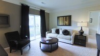 This single-floor space centers around a thoughtfully designed living room that balances comfort and style. A large sliding glass door serves as a prominent architectural feature, allowing generous natural light to fill the room while providing access to an outdoor balcony area with exterior views. Flanking the door are floor-to-ceiling black drapes that add a bold, dramatic contrast to the neutral, light-toned walls and create an elegant visual anchor.

The living room is finished with plush wall-to-wall carpeting in a warm beige tone, contributing to a cozy and inviting atmosphere. Furnishings include a cream-colored sofa accented with dark throw pillows, complemented by a matching dark armchair positioned near the window. A round, dark wood coffee table serves as the centerpiece of the seating arrangement, adding warmth and a sense of sophistication. A framed artwork featuring earthy gold and brown tones is mounted on the wall, tying together the room's cohesive color palette. Crown molding along the ceiling adds a refined architectural detail, enhancing the overall polished and well-finished character of the space.
