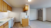 Located at 186 Mill St in Athens, Ohio, this single-floor unit offers a practical and straightforward layout designed for comfortable everyday living. The kitchen is equipped with warm honey-toned wood cabinetry, light countertops, a white electric range with coil burners, a dishwasher, a full-size refrigerator, and a microwave. The kitchen features easy-to-clean vinyl plank flooring and painted concrete block walls, giving it a durable, low-maintenance character. Adjacent to the kitchen, the open living area transitions to soft carpeting, providing a comfortable and versatile space. A window allows natural light to brighten the room, while a ventilation unit ensures climate control. The bedroom area continues the carpeted flooring and includes a generously sized closet with a full-width hanging rod and shelving, offering excellent storage capacity. A built-in desk area is accessible nearby, making the space well-suited for study or work. The bathroom features large-format white tile walls, a toilet, and vinyl plank flooring, maintaining a clean and simple aesthetic throughout. The unit's consistent use of neutral tones and practical finishes creates a cohesive, functional living environment well-suited for those seeking straightforward, low-maintenance accommodations in Athens.