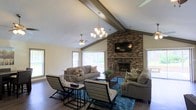 Located at 167 Briarwood Dr. in Jackson, Mississippi, this single-story home offers a welcoming and well-appointed interior designed for comfortable living. The standout feature of the main living area is its soaring vaulted ceiling adorned with exposed wood beams, which lend a warm, rustic character to the space. A full-height brick fireplace serves as the room's focal point, complemented by a mounted flat-screen television above the mantle, making it both a cozy and functional gathering space. Rich dark hardwood flooring runs throughout, providing an elegant contrast to the light-toned walls. Natural light pours in through large windows dressed with vertical blinds, offering views of the surrounding outdoor area. The living room is generously sized, accommodating multiple seating arrangements with ease, and is illuminated by a classic chandelier and a ceiling fan that add to the room's layered lighting. The overall ambiance strikes a balance between casual comfort and refined style, with thoughtful finishes and architectural details that elevate the everyday living experience. This home's single-floor layout ensures seamless flow and accessibility throughout the space.