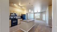 Located at 9701 Pearl St in Thornton, CO, this well-appointed single-floor residence offers two bedrooms and two full bathrooms within a thoughtfully designed layout. The open-concept kitchen serves as the heart of the home, featuring light maple cabinetry, dark granite-style countertops, a center island with a built-in sink, pendant lighting, and black appliances including a range, microwave, and refrigerator. Wood-style luxury flooring flows throughout the kitchen and living areas, while plush carpet adds comfort to the bedrooms. The spacious primary bedroom includes a ceiling fan, natural light from a large window with outdoor views, and direct access to its en-suite bathroom. The en-suite bath is equipped with a granite-topped vanity, wood cabinetry, a glass-enclosed shower, and a walk-in closet with wire shelving. The second bedroom also features a ceiling fan, a well-sized window with outdoor views, and its own adjacent full bathroom with matching finishes and a dedicated walk-in closet. Both bathrooms share a consistent design aesthetic with warm neutral tones and wood-style flooring. The overall color palette throughout is composed of warm beige and taupe tones, creating a cohesive and inviting atmosphere.