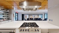 Located at 1200 North Veitch Street in Arlington, VA, this thoughtfully designed single-floor communal space offers a generous, open-concept layout ideal for socializing, dining, and relaxed gatherings. The space seamlessly blends several distinct functional zones within a bright, contemporary interior.

At its heart is a professional-grade kitchen anchored by a stainless steel Viking rangetop and a matching Viking range hood, set above a sleek white island with bar seating. Adjacent to the kitchen, a dedicated dining area features multiple tables and modern chairs beneath warm wood-paneled ceiling accents and pendant lighting, creating an inviting atmosphere for shared meals.

Large windows throughout the space invite abundant natural light and frame lush greenery views, enhancing the open, airy feel. The lounge area offers plush seating — including a substantial sectional sofa and bold orange accent chairs — arranged around a coffee table under geometric pendant fixtures, with a mounted flat-screen television adding to its comfort.

The entry area features large glass doors, terrazzo tile flooring, and decorative art panels, establishing an impressive first impression. Slatted wood privacy screens and built-in display shelving add architectural interest throughout. The cohesive design palette of white, wood tones, and contemporary furnishings gives the entire space a polished, modern character well-suited for a vibrant residential or professional community.