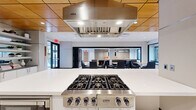 Located at 1200 North Veitch Street in Arlington, VA, this thoughtfully designed single-floor communal space offers a generous, open-concept layout ideal for socializing, dining, and relaxed gatherings. The space seamlessly blends several distinct functional zones within a bright, contemporary interior.

At its heart is a professional-grade kitchen anchored by a stainless steel Viking rangetop and a matching Viking range hood, set above a sleek white island with bar seating. Adjacent to the kitchen, a dedicated dining area features multiple tables and modern chairs beneath warm wood-paneled ceiling accents and pendant lighting, creating an inviting atmosphere for shared meals.

Large windows throughout the space invite abundant natural light and frame lush greenery views, enhancing the open, airy feel. The lounge area offers plush seating — including a substantial sectional sofa and bold orange accent chairs — arranged around a coffee table under geometric pendant fixtures, with a mounted flat-screen television adding to its comfort.

The entry area features large glass doors, terrazzo tile flooring, and decorative art panels, establishing an impressive first impression. Slatted wood privacy screens and built-in display shelving add architectural interest throughout. The cohesive design palette of white, wood tones, and contemporary furnishings gives the entire space a polished, modern character well-suited for a vibrant residential or professional community.