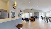 This single-floor community space is designed with an open-concept layout that seamlessly connects the kitchen, lounge, and dining areas. The soaring vaulted ceilings, recessed lighting, and light hardwood-style flooring throughout lend the space a bright and airy atmosphere.

The fully equipped kitchen is a standout feature, boasting stainless steel appliances, a mosaic tile backsplash, warm wood-toned cabinetry, and light quartz countertops. A peninsula with bar-height seating and stylish pendant lighting makes it ideal for casual entertaining. Adjacent to the kitchen, a high-top dining area with industrial-style metal chairs and large windows offers abundant natural light and exterior views.

The lounge area is generously proportioned, centered around a dramatic floor-to-ceiling stone fireplace that serves as the room's focal point. Comfortable leather sectional seating, mid-century modern accent chairs, and a wall-mounted television create a welcoming and functional gathering space. A ceiling fan adds to the comfort of the room, which also benefits from French doors leading to an outdoor area. Decorative wall art and eclectic furnishings give the space a warm, lived-in character. A separate fitness room with glass French doors is also accessible directly from the main area.