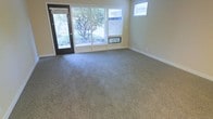 This single-story space features a open living area finished with wall-to-wall neutral-toned carpet that adds warmth and comfort underfoot. The room is bathed in natural light, drawn in through a large multi-panel window along the front wall and a glass-panel entry door that frames a view of mature trees and green landscaping outside. An additional smaller window positioned higher on an adjacent wall further brightens the interior. The walls are finished in a soft, warm beige tone that complements the understated color palette throughout. A wall-mounted HVAC unit sits beneath the large front window, providing climate control directly within the living space. White baseboards provide a clean contrast against the carpeted floors and painted walls. The layout is straightforward and functional, offering an open, flexible floor plan well-suited for various furniture arrangements. The overall ambiance is calm and understated, with an emphasis on natural light and a connection to the outdoor surroundings visible through the generous window placement.
