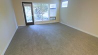 This single-story space features a open living area finished with wall-to-wall neutral-toned carpet that adds warmth and comfort underfoot. The room is bathed in natural light, drawn in through a large multi-panel window along the front wall and a glass-panel entry door that frames a view of mature trees and green landscaping outside. An additional smaller window positioned higher on an adjacent wall further brightens the interior. The walls are finished in a soft, warm beige tone that complements the understated color palette throughout. A wall-mounted HVAC unit sits beneath the large front window, providing climate control directly within the living space. White baseboards provide a clean contrast against the carpeted floors and painted walls. The layout is straightforward and functional, offering an open, flexible floor plan well-suited for various furniture arrangements. The overall ambiance is calm and understated, with an emphasis on natural light and a connection to the outdoor surroundings visible through the generous window placement.