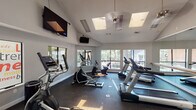 Located at 2642 Verandah Ln in Arlington, TX, this fitness center is a well-appointed, single-floor facility designed to support a comprehensive workout experience. The main exercise area features soaring vaulted ceilings that create an open, airy atmosphere, complemented by multiple skylights and large windows that flood the space with abundant natural light and offer views of the surrounding tree-lined exterior. The dark rubber flooring throughout provides both durability and traction underfoot.

Cardio equipment is a central focus, with multiple treadmills and elliptical machines thoughtfully arranged across the main floor. Full-length wall mirrors expand the sense of space and allow users to monitor their form during workouts. Multiple wall-mounted flat-screen televisions are positioned throughout the room, ensuring visibility from virtually every piece of equipment.

Two dedicated side rooms extend the facility's functionality. One room serves as a cycling studio, equipped with several stationary bikes and mirrored walls. The adjacent room provides a designated area for free weight training, including dumbbells and additional equipment. Motivational fitness-themed signage decorates the windows and walls, reinforcing the energetic ambiance of the space. Ceiling fans help maintain comfortable airflow throughout the facility.