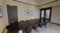 This single-floor conference room offers a functional and professional environment well-suited for meetings, presentations, or collaborative work sessions. The room is anchored by a large, dark espresso-toned conference table that comfortably seats numerous attendees, surrounded by matching dark wood and chrome chairs that lend a cohesive, polished aesthetic throughout the space.

The walls are finished in a neutral warm beige tone, complemented by dark wood crown molding that adds a refined architectural detail. Two framed black-and-white photographs depicting notable civic or governmental buildings are displayed on the main wall, contributing a classic and distinguished ambiance to the room.

Entry to the space is provided through a frosted glass-paneled door, allowing diffused light to filter in while maintaining privacy. A secondary solid door is also present, offering additional access or utility. The room is equipped with a clearly visible emergency exit sign, underscoring its compliance with safety standards. The light-toned flooring provides a subtle contrast to the dark furnishings, creating a balanced and professional atmosphere throughout.