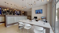 This single-floor lobby and mail center is designed with a clean, contemporary aesthetic that balances functionality with inviting style. Light hardwood flooring runs throughout the space, complementing the neutral warm tones of the walls and contributing to an open, airy atmosphere. Large floor-to-ceiling windows along one side of the room flood the interior with natural light while offering views of the surrounding outdoor greenery.

A central feature of the space is a substantial island with a crisp white quartz surface and dark wood base, surrounded by modern white molded chairs. This island serves as both a gathering point and a casual workspace. Adjacent to the island, a dedicated workstation area is equipped with desktop computers, providing a practical resource hub.

A wall-mounted flat-screen television is positioned above a textured stone accent wall, adding a media focal point to the room. Overhead, an eclectic collection of geometric and industrial-style pendant lights creates visual interest and warm ambient lighting.

The mail center is prominently integrated into the space, featuring an extensive bank of individual metal mailboxes set against a textured backdrop. Above the mailboxes, open shelving units with vibrant blue interiors display decorative objects, adding a pop of color. Dark wood-paneled architectural columns define zones within the space, with a glimpse into an adjacent lounge area visible beyond.