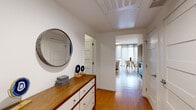 Located at 75 Brainerd Rd in Boston's Allston neighborhood, this model studio unit is a well-appointed single-floor space that makes efficient use of its layout through smart design and modern finishes. Warm hardwood flooring flows throughout, unifying the open-plan living area, dining space, and sleeping quarters into one cohesive environment. The living area is anchored by large floor-to-ceiling windows dressed with sheer drapes, allowing abundant natural light to fill the room while offering views of the surrounding cityscape. A compact, fully equipped kitchen features dark cabinetry, stainless steel appliances, and clean countertops, seamlessly integrated into the open floor plan. The hallway entrance is welcoming and functional, featuring a built-in wood-topped dresser with white cabinetry and a round decorative mirror — setting an inviting tone upon entry. The bathroom is finished in a clean, minimalist style, with a sleek undermount sink, light wood vanity, large wall mirror, and dark tile flooring, complemented by crisp white five-panel doors throughout. The overall ambiance is contemporary and bright, with a neutral color palette accented by bold artwork and vibrant décor, reflecting a stylish and livable modern urban aesthetic.