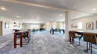 Located at 3571 Heritage Towne Blvd in Midlothian, Texas, this well-appointed game room is a generously sized single-floor recreational facility designed for social engagement and entertainment. The open-plan layout is anchored by stylish, mixed-tone carpet flooring that adds warmth and texture throughout the space.

The room is outfitted with an impressive array of entertainment options, including a pool table with a sleek dark frame and blue felt surface, a classic wooden shuffleboard table, vintage arcade cabinets, and a ping-pong table. Multiple seating arrangements — featuring bar-height tables with upholstered chairs, round tables for group gatherings, and bench seating — are thoughtfully distributed to encourage both active play and casual socializing.

A wall-mounted flat-screen TV is paired with bar seating, creating a media-focused lounge area, while pendant lighting overhead adds a warm, inviting ambiance. A dedicated kitchenette with a sink and open shelving supports hosting needs. Natural light pours in through multiple windows positioned around the room, and a set of French doors leads out to the balcony, offering a seamless indoor-outdoor connection. Recessed ceiling lighting throughout ensures the space remains bright and welcoming at all hours. The neutral, crisp white walls provide a clean backdrop that keeps the focus on the vibrant amenities within.