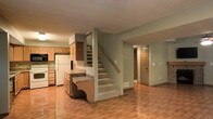 This two-story residential space offers a practical and welcoming layout across both levels. The main floor showcases an open-concept design where the kitchen flows naturally into the living area, creating a connected and versatile living environment. The kitchen is equipped with warm-toned wood cabinetry, a white refrigerator, a built-in microwave, and a peninsula countertop that provides additional workspace and casual seating potential. The countertops feature a stone-look finish that complements the overall color palette. Light wood-style flooring runs throughout the main level, adding warmth and continuity to the space. The walls are finished in a soft, neutral gray tone, contributing to a calm and understated ambiance. A textured ceiling adds a classic residential character throughout. A carpeted staircase with a wooden handrail leads to the upper floor, providing a smooth transition between levels. The layout also includes a doorway leading to additional rooms or utility spaces, enhancing the overall functionality of the home. The space is well-suited for everyday living, with a straightforward floor plan that balances openness with defined areas for cooking, dining, and relaxing.