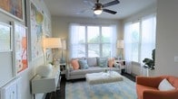 This single-floor space showcases a thoughtfully designed living area that blends comfort and style in a light-filled, coastal-inspired setting. The living room is the centerpiece of the space, featuring large windows dressed with sheer white curtains and horizontal blinds that allow generous natural light to filter in while offering views of the surrounding greenery outdoors. A ceiling fan with an integrated light fixture is centrally mounted, providing both function and a clean architectural detail overhead. The room is anchored by a plush grey sofa adorned with soft coral and light blue accent pillows, creating a relaxed yet polished color palette. A textured white ottoman serves as a coffee table, complemented by a clear acrylic tray and decorative accessories. A patterned chevron area rug in blue and white grounds the seating arrangement, adding visual interest to the dark hardwood flooring. Flanking table lamps with white shades and blue ceramic bases provide warm ambient lighting. A white console or sideboard along one wall offers display and storage space, decorated with greenery and sculptural accents. Artwork on the walls adds a creative touch, tying the coastal theme together throughout this inviting, well-appointed living space.