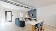 Located at 1301 Mulberry St in the heart of Des Moines, Iowa, this contemporary single-floor common space offers a sophisticated blend of modern design and functional amenities across a generously sized footprint. The open-plan layout seamlessly connects the kitchen and living areas, creating an inviting, versatile environment.

The kitchen is outfitted with stainless steel appliances, dark navy cabinetry, a crisp white countertop, and a fluted-panel island with bar seating in bold teal. Sculptural gold globe pendant lights hang above, complemented by warm wood-slat wall paneling that adds texture and depth.

The living area features exposed ductwork that lends an industrial edge to the otherwise refined interior. A dramatic dark charcoal built-in media wall serves as a focal point, while light wood-look flooring and large windows allow natural light to fill the space. An outdoor balcony offers urban views of the Des Moines skyline.

The accessible restroom is thoughtfully designed with warm wood wainscoting, bold geometric patterned wallpaper, globe sconce lighting, and an arched mirror, delivering a boutique aesthetic. A spacious courtyard with tiled pavers and outdoor seating rounds out this well-appointed property.