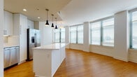 This single-floor residence offers a clean, contemporary design with a cohesive aesthetic carried throughout every room. The heart of the space is an expansive open-concept kitchen and living area, where warm hardwood flooring stretches across the full layout. The kitchen is outfitted with sleek white shaker cabinetry, a large central island with a crisp white countertop, stainless steel appliances including a gas range, microwave, and refrigerator, and stylish pendant lighting that adds a modern accent. Recessed lighting and under-cabinet illumination further enhance the kitchen's functionality. The adjoining living area is bathed in natural light from large windows dressed with horizontal blinds, offering views of the surrounding cityscape. The bedrooms continue the warm hardwood flooring theme, featuring clean white walls, ample closet space, and windows that bring in additional natural light. The bathrooms are finished with light ceramic tile flooring, white shaker-style vanities topped with granite countertops, and large mirrors. The primary bathroom features a double-sink vanity and a tiled shower. Throughout the residence, the bright white palette, quality finishes, and abundant natural light create a welcoming and airy atmosphere.