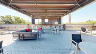 Located at 1235 Water Tower Cir in Greenville, SC, this generously sized rooftop terrace offers a compelling blend of industrial design and open-air leisure. The space is defined by its striking corrugated metal overhead canopy, supported by exposed steel beams, which shelters a substantial portion of the terrace while still allowing abundant natural light and fresh air to flow through. The flooring throughout is composed of durable paver brickwork, lending a clean, urban aesthetic underfoot. Beneath the covered section, a built-in bar with high-top seating provides a dedicated social hub, complemented by multiple lounge seating arrangements featuring modern sofas, accent chairs, and bistro tables — ideal for both relaxed gatherings and more structured social events. The open-air portion of the terrace extends the usable area significantly, furnished with additional lounge chairs, fire pit seating, and picnic-style tables. Corrugated metal siding lines the perimeter walls and utility structures, reinforcing the contemporary industrial character of the space. Panoramic views of the surrounding Greenville skyline and lush tree canopy are visible from multiple vantage points, enhancing the overall ambiance. The Water Tower landmark is also visible, adding a notable sense of place to this well-appointed outdoor venue.