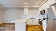 This well-designed 2-bedroom, 1-bathroom unit offers a clean, modern aesthetic across a single floor. The open-concept kitchen and living area serves as the heart of the space, featuring crisp white cabinetry, a subway tile backsplash, quartz countertops, a kitchen island, and a full suite of stainless steel appliances including a refrigerator, electric range, and built-in microwave. Warm, wide-plank hardwood-style flooring runs throughout the entire unit, lending a cohesive and inviting feel to every room. The living area is bright and airy, benefiting from a large window that frames views of the surrounding neighborhood and hillside. A private balcony with glass-panel railings extends the living space outdoors, offering an elevated view of the landscaped courtyard and community buildings. The two bedrooms are comfortable and neutrally finished, with clean white walls and continued hardwood-style flooring. A dedicated closet provides additional storage. The bathroom is thoughtfully appointed with a white soaker tub and shower combo, a white vanity with grey countertop, a large mirror, and recessed lighting. Two hallways connect the rooms efficiently, completing this well-organized and contemporary layout.