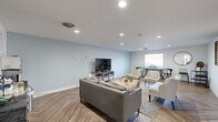 This thoughtfully designed living room offers a cohesive and modern aesthetic across its single-floor layout. The space is defined by striking herringbone-patterned wood-look flooring that runs throughout, adding visual depth and a sophisticated touch to the overall design. Soft blue-gray walls create a calm, contemporary atmosphere, complemented by crisp white trim and a smooth white ceiling fitted with recessed lighting that evenly illuminates the entire room.

A large picture window is a standout architectural feature, flooding the space with natural light and framing an expansive scenic view — ideal for both relaxation and productivity. The room is arranged to accommodate multiple functional zones: a comfortable seating area centered around a distinctive live-edge wood coffee table, paired with a gray sofa, mid-century modern accent chairs, and a loveseat, all anchored by a patterned area rug. Decorative wall art in geometric and organic motifs adds character to the white-painted accent wall.

A media console with a wall-mounted flat-screen television serves as a focal point on one end, while a work or dining area near the window features a metal-frame table and matching chairs. A door along one wall provides additional access to the exterior. The overall ambiance is clean, airy, and inviting — balancing modern design with warm, natural elements.