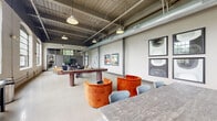 Located at 3201 First Ave in Columbus, GA, this single-floor amenity space offers a generously proportioned open-plan layout that blends industrial character with modern comfort. The architecture showcases exposed structural beams, painted brick walls, and polished concrete floors, all topped by a soaring ceiling with visible ductwork and pendant lighting that gives the space an authentic loft-like atmosphere.

Large, arched multi-pane windows line the walls, flooding the interior with abundant natural light and offering views of the surrounding greenery. The open floor plan is thoughtfully organized into distinct zones. A recreation area houses both a shuffleboard table and a billiard table, set atop striped area rugs. A lounge zone features plush leather wingback chairs and bold orange velvet seating, creating inviting gathering spots. A dining and collaboration area offers communal tables with mixed seating, suitable for casual meetings or social events.

Curated wall art, including vibrant portrait pieces and abstract compositions, adds a gallery-like quality throughout. A glass-partitioned section provides a more enclosed lounge experience with a television and additional seating. The overall ambiance is dynamic yet welcoming, balancing the rawness of the industrial architecture with warm, contemporary furnishings and thoughtful design details.