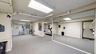 Located at 1189 Washington St in Middletown, Connecticut, this fitness facility spans a single floor and is divided into two functional exercise rooms designed to support a well-rounded workout experience. The space features neutral-toned concrete block walls, durable gray carpet flooring, and flush panel ceiling lights that provide consistent, even illumination throughout.

The primary exercise room is equipped with a variety of cardio machines, including treadmills, an elliptical, and a stationary bike, along with a multi-station strength training machine. Motivational fitness posters and posted exercise room rules are displayed on the walls, contributing to a focused workout environment. A wall-mounted television is also present, offering entertainment during cardio sessions.

The second room offers a more open layout, highlighted by a full wall of mirrors that enhance the sense of space and support activities such as stretching, mat work, or group exercise. Exposed overhead piping and recessed panel lighting give the area a straightforward, utilitarian character. The open floor plan provides flexibility for a range of fitness activities.

Together, the two rooms offer a compact yet comprehensive fitness facility suited for cardiovascular training, strength building, and open-floor exercise, making it a practical and well-equipped space for regular physical activity.