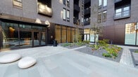 Second + Second is a contemporary residential building distinguished by its striking multi-story dark brick façade and thoughtfully designed ground-floor common spaces. The exterior courtyard sets a refined tone with large-format stone pavers, curated landscaping featuring ornamental grasses, shrubs, and slender young trees set against decorative gravel beds, and warm wood slatted panels that soften the building's bold, angular architecture.

Entry is through sleek black-framed glass double doors, opening into a well-appointed lobby that immediately impresses with its open layout and design-forward aesthetic. Warm wood slat ceiling panels extend throughout the interior, complementing the polished concrete flooring and creating an inviting, upscale atmosphere. A central reception desk anchors the space, surrounded by contemporary seating including teal accent chairs and sculptural lounge pieces. Gallery-style wall art adds a curated, residential-hotel quality to the environment.

Glass partitions divide functional zones within the lobby while maintaining a sense of openness and allowing natural light to flow freely throughout. A secondary vestibule connects the interior to the courtyard, featuring floor-to-ceiling glazing that frames views of the landscaped outdoor space. Building systems and access controls are cleanly integrated into the entryway walls, reflecting the space's attention to both form and function.