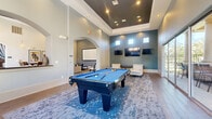 This well-appointed amenity and entertainment space offers a generously sized, single-floor layout designed for recreation and social gatherings. The main game room serves as the centerpiece, boasting soaring tray ceilings with a dramatic dark finish and recessed lighting that highlight the full-size billiards table below. A bold teal accent wall displays multiple mounted flat-screen TVs, while a shuffleboard table and comfortable lounge seating invite relaxed socializing. Large windows along one wall frame outdoor courtyard views and flood the space with natural light.

A dedicated golf simulator room provides an immersive experience with a Full Swing simulator screen, synthetic turf hitting mat, and a full rack of golf clubs — ideal for year-round play. Adjacent to the main lounge is a cozy screening nook, featuring a large projection screen, plush seating, and cinema-themed artwork that creates a private, theater-like atmosphere.

A café-style bar area with a dark granite countertop, mosaic tile backsplash, and pendant lighting anchors a social hub, complemented by a dining area with direct access to an outdoor space. Throughout, hardwood-style flooring, teal and gray tones, and arched architectural details tie each area together into a cohesive, modern design.