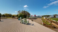 Located at 2060 Quingate Place in Halifax, Nova Scotia, this outdoor terrace space offers a well-appointed communal environment designed for relaxation and community engagement. The centerpiece is a generously sized composite wood deck, furnished with colourful Adirondack chairs and barbecue grills, providing a welcoming gathering area beneath open skies. The deck connects seamlessly to surrounding gravel pathways that wind through the space, leading residents toward multiple raised garden beds where seasonal plantings and community gardening thrive. Expansive, well-maintained green lawns frame the property on multiple sides, offering open recreational space and a refreshing contrast to the urban surroundings. The grounds are bordered by black iron fencing, providing a sense of enclosure while maintaining open sightlines to the surrounding Halifax neighbourhood. A modern multi-storey residential tower with a distinctive geometric panelled façade anchors the site, while additional residential buildings are visible in the backdrop, reinforcing the community-oriented character of the location. Mature trees and shrubs provide natural screening and shade throughout the terrace, contributing to a pleasant and park-like atmosphere. The space benefits from abundant natural light and expansive open sky views, making it a functional and inviting outdoor amenity year-round.