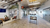This single-floor commercial space blends industrial heritage with contemporary design across a generously proportioned open layout. The main corridor serves as the central spine of the space, characterized by striking exposed wooden beam ceilings that reveal the building's historic structure alongside modern fluorescent strip lighting. Polished concrete floors run throughout, lending the space a raw yet refined aesthetic.

The entrance area features a welcoming lobby with display cases set atop a traditional area rug, offering an artful contrast to the industrial surroundings. A glass-fronted leasing office is conveniently positioned along the main corridor, furnished with modern seating and visible artwork, providing a professional and accessible point of contact.

A dedicated mail and package room lines both walls with extensive banks of black individual mailboxes and parcel lockers, complemented by a communal table and seating — a practical and well-organized amenity. Natural light filters in through large windows at the far end of this corridor, enhancing the open feel of the space.

Dual stainless steel elevators are situated in a sage-green alcove, providing accessible vertical circulation. Structural concrete columns punctuate the space throughout, reinforcing the building's industrial character. The overall ambiance is dynamic and purposeful, balancing utilitarian functionality with thoughtful, urban design details.