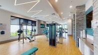 This single-level fitness center is designed with a contemporary aesthetic, offering a spacious and well-organized layout that accommodates a wide variety of workout disciplines. The main exercise floor features Matrix-brand strength and cardio equipment, including cable machines, weight benches, ellipticals, rowing machines, and treadmills, all arranged across warm hardwood-style flooring that extends throughout the space.

A defining architectural feature is the striking geometric LED lighting system installed across the high white ceilings, with angular, zigzag-pattern linear fixtures that lend a dynamic, modern energy to the environment. Large black-framed windows along the perimeter allow abundant natural light and provide views of lush outdoor greenery. Stacked stone columns and accent walls add a textural contrast to the otherwise clean, neutral palette.

A dedicated spin studio is separated by floor-to-ceiling glass walls, featuring deep navy blue walls, a wall-mounted television, recessed ceiling lighting with warm LED trim, and multiple cycling bikes. The main area also includes wall-mounted flat-screen televisions and decorative teal-toned art panels. Dual water fountains are conveniently positioned near the stacked stone columns, completing this thoughtfully designed and fully equipped fitness facility.