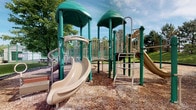Located on Chatman Drive in Strongsville, Ohio, this outdoor recreational space offers a well-equipped playground designed to encourage active play in a natural setting. The centerpiece of the space is a multi-tower play structure finished in green and tan, featuring dual dome-topped towers connected by walkways and panels. Children can enjoy a variety of play options, including a curved slide, a straight slide, climbing ladders, and interactive panel elements integrated throughout the structure. The entire play area is surfaced with a generous layer of wood chip mulch, providing a cushioned, safety-conscious ground cover beneath and around all equipment. The space is set within a verdant landscape, with mature trees framing the perimeter and offering natural shade. Open sky and rolling greenery in the background contribute to a bright, welcoming outdoor atmosphere. The layout of the playground allows for multiple children to engage simultaneously across different activity zones, making it a functional and engaging community recreation destination.