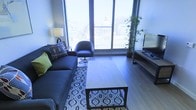 This single-floor urban living space offers a thoughtfully designed interior with a strong connection to the city skyline beyond. The living area is anchored by large floor-to-ceiling windows that flood the room with natural light and frame impressive views of the surrounding urban landscape, including notable architectural landmarks visible in the distance.

The living room features light hardwood flooring that complements the contemporary aesthetic throughout. A deep navy sofa adorned with decorative accent pillows provides comfortable seating and faces a sleek media console housing a flat-screen television. A wooden coffee table sits atop a patterned area rug, adding visual texture and warmth to the space. A modern accent chair in a matching dark tone completes the seating arrangement.

A tall bamboo plant positioned near the windows introduces a natural element, softening the urban atmosphere. Ambient lighting is provided by a traditional table lamp, offering a warm counterpoint to the cool tones of the space. The overall ambiance is contemporary and polished, blending city energy with comfortable, well-considered interior design.