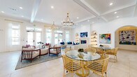 This sophisticated single-floor community space showcases a coastal-inspired design with a bright, airy palette of crisp whites, warm wood accents, and vibrant teal, navy, and magenta furnishings throughout. The expansive main lounge area features coffered ceilings adorned with gold-toned chandelier lighting and large French doors that flood the space with natural light. A stylish seating area anchors one end of the lounge, centered around a fireplace flanked by illuminated built-in arched shelving and a wall-mounted television, creating an inviting focal point. A glass-top dining table with woven rattan chairs adds a casual yet refined dining area within the open-plan layout. A separate room with soaring vaulted ceilings clad in natural wood showcases a wooden dining table surrounded by bold fuchsia upholstered chairs, built-in cabinetry with cubbies, and dramatic teal drapery. The welcoming entry foyer features large-format tile flooring, arched architectural details, and eclectic sculptural seating elements. Throughout the space, carefully curated artwork, arched doorways, and a harmonious blend of textures—woven, velvet, and natural wood—create a warm, upscale ambiance ideal for gathering and socializing.