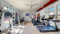 This fitness center is a comprehensively outfitted single-floor facility designed to accommodate a full range of workout needs. The space is bright and open, with large windows lining the walls and allowing abundant natural light to stream in throughout the day, offering views of the surrounding outdoor greenery. Light wood-style flooring runs throughout, lending a clean and modern aesthetic to the interior.

The cardio zone is prominently featured, housing multiple treadmills and elliptical machines from Life Fitness, as well as stationary and cycling bikes. A wall-mounted flat-screen television is positioned for easy viewing during cardio sessions, and ceiling fans overhead contribute to comfortable air circulation. A distinctive red ventilation duct runs along the upper portion of one wall, adding an industrial design accent to the otherwise crisp, white interior.

The strength training area includes a cable machine, a multi-function rack loaded with weight plates, a dedicated dumbbell rack with a full range of free weights, and adjustable benches. Medicine balls and stability equipment are also available. Colorful artwork on the walls adds visual interest. The space is well-lit, well-ventilated, and thoughtfully organized to support both cardio and resistance training workouts.