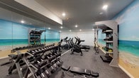 This fitness center is a thoughtfully designed, single-level space that provides a comprehensive workout environment. Its most striking design element is a vivid, floor-to-ceiling beach-scene mural that spans the length of one wall, depicting a tropical shoreline complete with turquoise waters, sandy beach, and a bright sky. The mural gives the space an energetic, resort-like atmosphere. A full-wall mirror runs along the opposite side of the room, visually expanding the space and enhancing the open, airy feel.

The flooring consists of durable grey rubber, suitable for both cardio and strength training. Recessed ceiling lighting provides bright, even illumination throughout. The cardio section is equipped with multiple Star Trac treadmills, elliptical trainers, and stationary cycling bikes. A multi-station cable machine is positioned toward the rear of the space for strength and resistance training.

Free weights are organized on tiered dumbbell racks, offering a broad range of weight options. A dedicated TRX suspension training station is mounted to a freestanding rig, which also includes shelving for medicine balls and accessories. An adjustable weight bench provides versatility for a wide range of exercises. Together, the equipment selection and design make this fitness center a well-rounded facility suited to varied training needs.