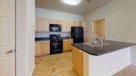 This single-floor unit offers a functional and well-organized layout across several distinct areas. The living space features neutral-toned carpet, smooth cream walls, and a ceiling fan with light fixture, creating a comfortable and adaptable environment. Natural light enters through blinded windows, and a glass-panel door provides direct outdoor access, adding brightness to the room.

The kitchen is equipped with light wood cabinetry, dark countertops, and black appliances including a range, microwave, and refrigerator. Light hardwood-style flooring runs throughout the kitchen, and a peninsula with a sink adds practical counter space and a natural divide between the kitchen and adjacent areas.

The bedroom is generously sized with plush carpeting, a ceiling fan, and a window allowing natural light. A walk-in closet with built-in shelving and rod offers ample storage. Two interior doors open directly to the full bathroom, which features tile flooring, a tub/shower combination, a vanity with wood cabinetry, and a large mirror.

A dedicated laundry room with hardwood-style flooring houses a full-size washer and dryer. The entry area transitions seamlessly from tile to carpet, completing this practical and cohesive living space.