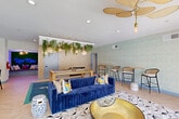 Located at 2151 Cumberland Pkwy SE in Atlanta, Georgia, this thoughtfully designed clubhouse amenity space offers a compelling blend of lively social areas and resort-style outdoor recreation. The single-floor interior is divided into distinct zones that cater to both relaxation and entertainment. The main lounge area is anchored by a plush, royal blue velvet sofa paired with a striking gold bowl coffee table and a geometric-patterned area rug, all beneath a sculptural gold leaf chandelier. A dramatic overhead trellis adorned with cascading greenery and woven pendant lights creates a lush, botanical atmosphere above a full-size billiards table. High-top bar seating with woven chairs lines one side, complemented by a wet bar with gray cabinetry and arched mirror accents. Floor-to-ceiling sliding glass doors open directly to the outdoor pool deck, seamlessly connecting the interior to the resort-style amenity space outside. A cozy built-in bench nook features a vivid moss wall backdrop with a neon "Uw Moss" sign, adding a playful, branded touch. The outdoor area showcases a generously sized swimming pool with a surrounding sun deck, lounge chairs, umbrellas, and lush tree-lined views, creating an inviting retreat within the Atlanta community.