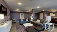 This single-floor space showcases a thoughtfully designed lounge area that combines bold aesthetics with comfortable, functional furnishings. The walls are painted in a deep, rich purple tone, creating a dramatic and inviting atmosphere that serves as a striking backdrop throughout the space. Recessed ceiling lights are evenly distributed across the smooth ceiling, providing warm, consistent illumination throughout the room.

The seating arrangement is both generous and well-organized, featuring multiple dark charcoal-gray sofas and accent chairs positioned around a sleek white coffee table with a modern acrylic base. A patterned armchair adds a decorative contrast to the neutral upholstery of the surrounding seating. Decorative throw pillows in geometric and textured patterns are layered across the furniture, contributing to the polished, curated look.

Anchoring the seating area is a vibrant, multi-colored area rug with an abstract burst pattern, introducing energy and visual interest against the dark hardwood-style flooring. A structural column near the center of the room integrates seamlessly into the design. Wall art and a table lamp with a white shade add finishing touches, while a white door is visible at the rear, suggesting additional space beyond.