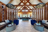 Located at 50 Woodview Way in Franklin, MA, this single-story clubhouse at Halstead Franklin is a boldly designed communal space that masterfully blends rustic architectural elements with vibrant, contemporary interiors. The soaring vaulted ceiling, supported by exposed wooden trusses, creates a dramatic sense of height and openness throughout the main living area. Underfoot, an intricate geometric parquet-style hardwood floor adds a refined elegance that contrasts beautifully with the space's eclectic character.

The living room is a visual standout, featuring rich, large-scale floral wallcoverings in deep jewel tones that energize every wall. Plush navy-blue velvet seating, pendant globe lighting, a central island with tiled base, and a tiled accent fireplace wall provide distinct gathering zones within the open floor plan. A dedicated game area offers a full golf simulator alongside a shuffleboard table, set on herringbone brick flooring for a warm, textural contrast.

Large windows and glass doors flood the interior with natural light while framing views of the outdoor pool area and surrounding greenery. The patio extends the living space outdoors, offering additional seating amid a lush landscape. Every detail of this clubhouse reflects a carefully curated, hospitality-forward design aesthetic.
