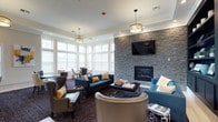 The clubhouse at Brookhaven Lofts is a thoughtfully designed single-floor communal space that blends contemporary style with warm, inviting elements. The layout flows seamlessly from a welcoming entryway—finished with light hardwood flooring, crown molding, and soft yellow walls—into a series of distinct yet connected social areas.

The main lounge serves as the centerpiece of the space, anchored by a striking floor-to-ceiling stacked stone accent wall housing a linear gas fireplace and a wall-mounted flat-screen television. Built-in dark shelving flanks the fireplace, offering decorative display space and adding depth to the room. Seating is plentiful, with tufted teal sofas, leather wingback chairs, and upholstered accent chairs arranged around a textured area rug, creating an atmosphere that is both sophisticated and comfortable.

An abundance of large windows lines the perimeter, flooding the space with natural light and offering views to the outdoors. A dedicated dining nook accommodates a round table with stylish upholstered chairs, ideal for casual gatherings. Pendant lighting with gold-toned drum shades provides warm illumination throughout. A wet bar area, visible from the entryway and framed by the same stacked stone detailing, adds a refined, amenity-rich finishing touch to this well-appointed communal retreat.
