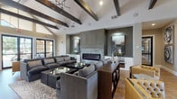 This single-story communal space combines rustic architectural charm with contemporary design, creating a welcoming and functional environment. The main lounge area is defined by soaring vaulted ceilings adorned with bold, dark wood beams that provide striking visual contrast against crisp white surfaces. A branch-style chandelier adds an artisanal touch overhead. The focal point of the lounge is a floor-to-ceiling stone fireplace framed by sage-green cabinetry with paneled millwork detailing, flanked by curated artwork. Generous sectional sofas in charcoal gray surround low-profile coffee tables, encouraging social gathering. Warm hardwood flooring flows throughout the space, complementing the neutral color palette. Large black-framed glass doors and tall windows along the perimeter invite abundant natural light and provide views to the outdoors. An adjacent kitchen and bar area features an expansive island with a slate-toned countertop and leather-upholstered barstools, white cabinetry, open floating shelves, pendant lighting, and a wall-mounted television—making it well-suited for casual entertaining and everyday use.